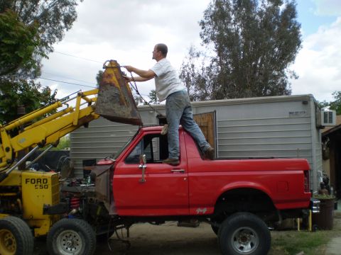 1969 Ford F-250 - A man stands on the roof of a red pickup truck while operating a yellow loader.