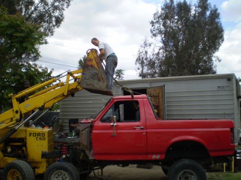1969 Ford F-250 - A red pickup truck is being loaded with dirt by a yellow backhoe, with a man standing on the backhoe's bucket.