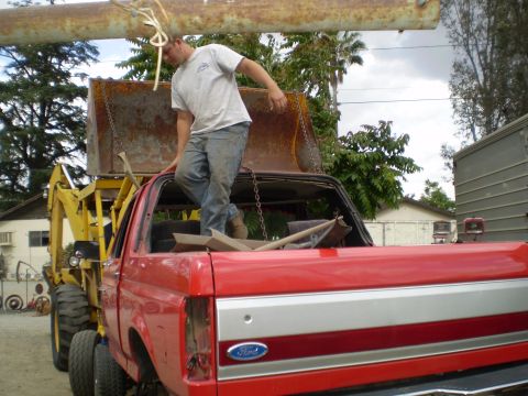1969 Ford F-250 - A red pickup truck, possibly from the 1980s, is being worked on as a backhoe lifts a large pipe above it.