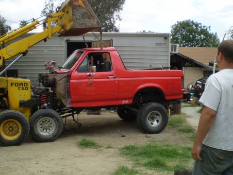 1969 Ford F-250 - A red pickup truck body is being lifted by a yellow backhoe loader.