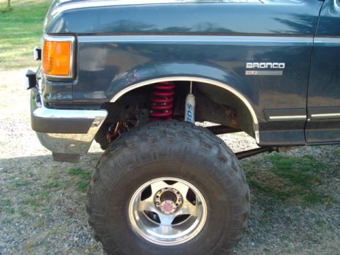 1990 Ford Bronco - Driver-side front wheel-well detail of a dark blue Ford Bronco with a 'Bronco XLT' fender badge, showing lifted front coil suspension with…