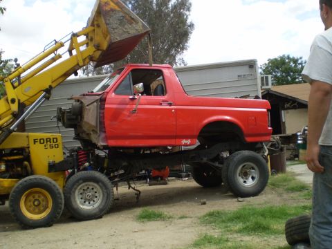1969 Ford F-250 - A red pickup truck is being lifted by a front loader, revealing its chassis and suspension components.
