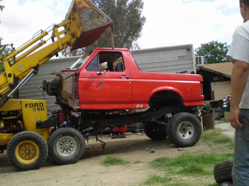 Red pickup truck body being lifted by a yellow Ford E50 front loader. The truck has a 4x4 decal