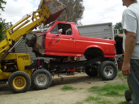 1969 Ford F-250 - A red Ford truck body is being lifted by a yellow loader, revealing its chassis and suspension system.