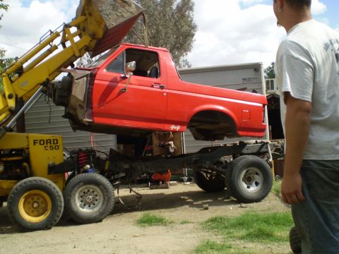 1969 Ford F-250 - A red pickup truck body is being lifted off its chassis by a yellow loader, with a man watching.