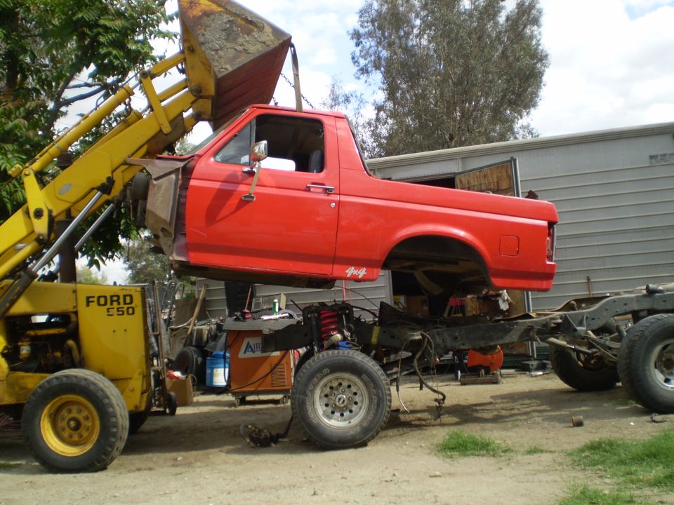 Red Ford pickup truck cab and bed lifted by yellow Ford E50 loader. Frame and chassis visible below