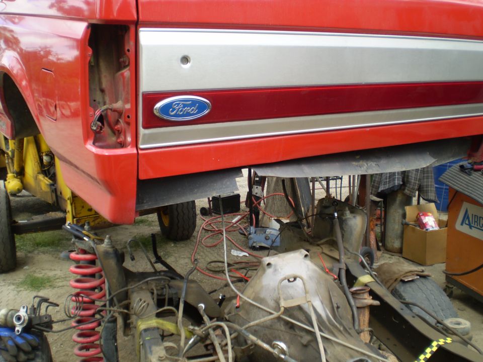Red Ford truck bed lifted, showing chassis and drivetrain components underneath. Ford logo visible
