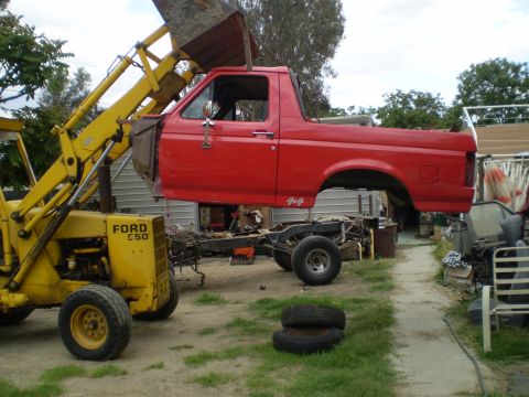 1969 Ford F-250 - A red pickup truck body is being lifted by a yellow loader in a yard with other parts visible.
