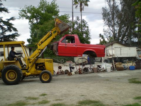 1969 Ford F-250 - A red pickup truck cab is being lifted by a yellow Ford loader, with other vehicles and parts visible in the background.