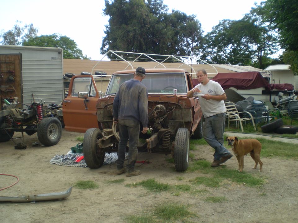 Orange Ford F-Series truck with front end removed, two men standing nearby, one holding a plate, a dog stands to the right