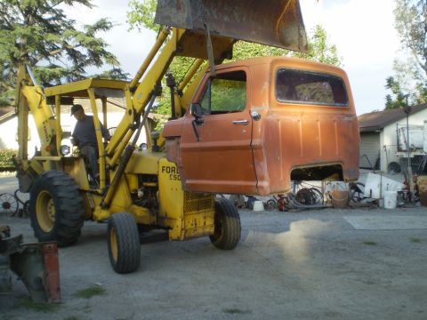 1969 Ford F-250 - A backhoe is lifting the cab of an orange Ford truck. The backhoe itself is yellow and has 'FORD E50' visible.