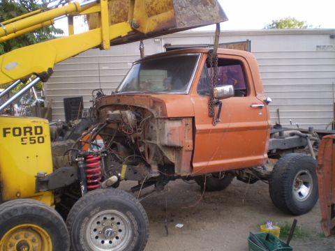 1969 Ford F-250 - A 1969 Ford F-250 truck cab is hoisted by a yellow loader, revealing its disassembled front end.