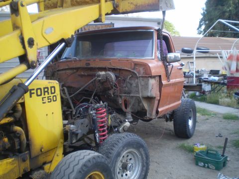 1969 Ford F-250 - A 1967 Ford truck's front end is suspended by a yellow loader, revealing its engine bay and suspension components.