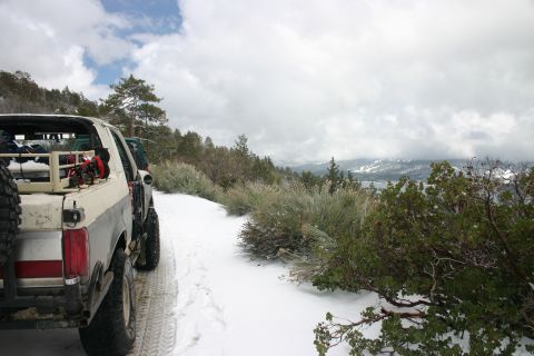 1991 Ford Bronco - Two SUVs are parked on a snow-covered road in a mountainous area with trees and cloudy skies.