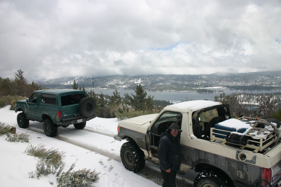 guy next to white pickup truck, green Bronco behind it, both in snow with lake and mountains in background