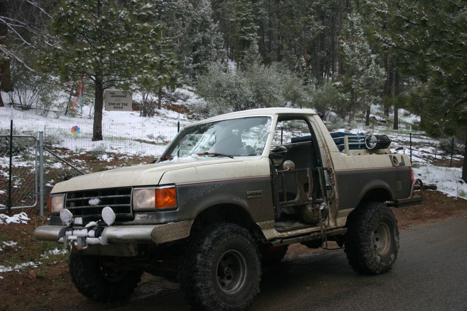 Ford Bronco with off-road modifications parked on a dirt road with snow on the ground and trees in the background. The driver's door is open