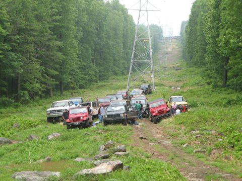 1985 Ford Bronco - A group of off-road vehicles are parked on a dirt path surrounded by trees, with large power line towers in the background.