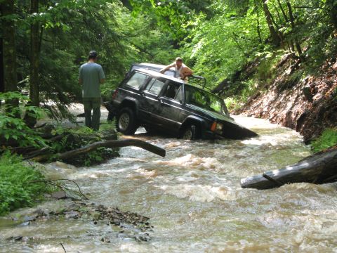 1985 Ford Bronco - A Jeep Cherokee XJ navigates a shallow, rocky stream in a wooded area.