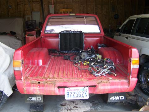 1988 Suzuki Samurai - Rear view of a red pickup truck bed containing a radiator and a tangled wiring harness. Another vehicle is partially visible to the right.