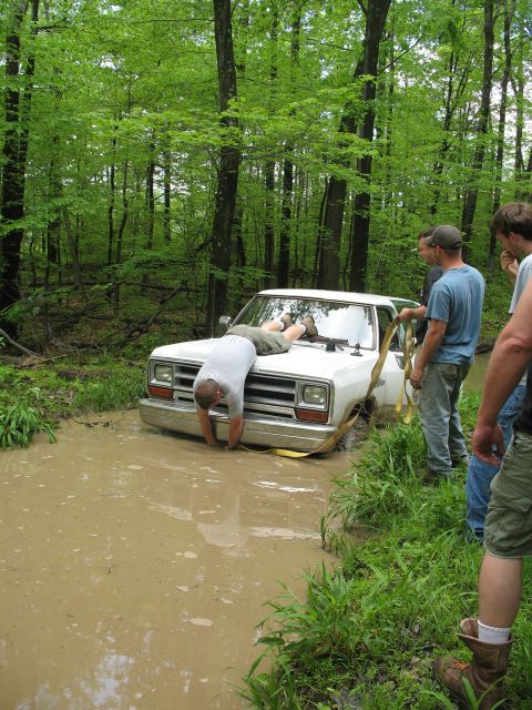1985 Ford Bronco - A group of people attempt to free a white pickup truck that is stuck in a muddy ditch in a wooded area.