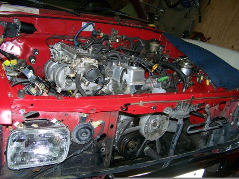 1988 Suzuki Samurai - The engine bay of a red vehicle is shown, with various engine components and wiring. The headlight is detached.