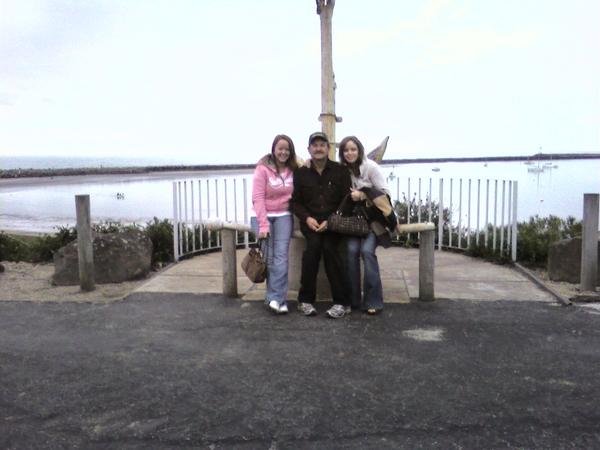 Three people stand on a paved area near a white railing overlooking a body of water with boats