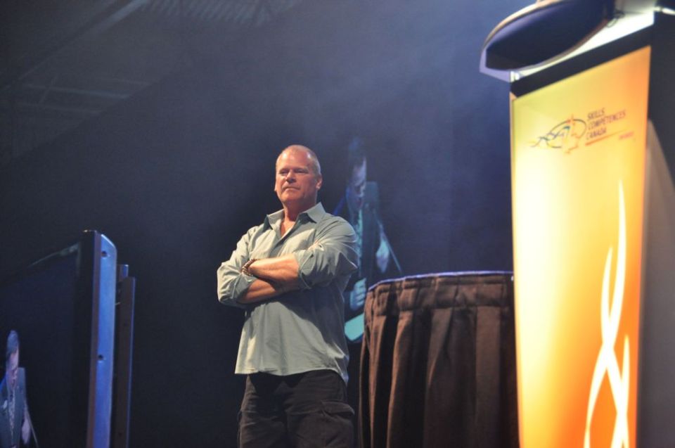 Man with arms crossed stands on a stage next to a podium with a banner