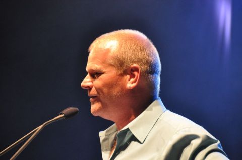 1996 Ford Bronco - A man in a light blue collared shirt speaks into a microphone on a dark blue background.