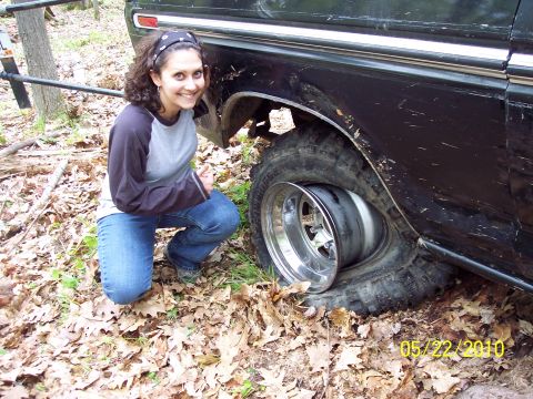 1979 Ford Bronco - A woman kneels beside a vehicle with a severely flat tire in a wooded area.