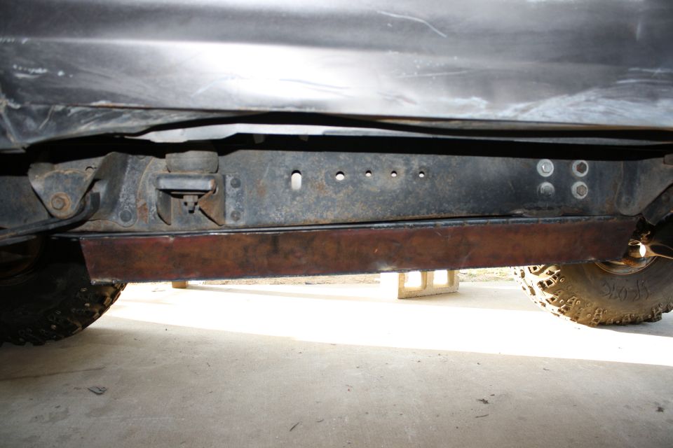 Underside of a vehicle showing the frame rails, suspension components, and a tire. Some rust is visible on the frame