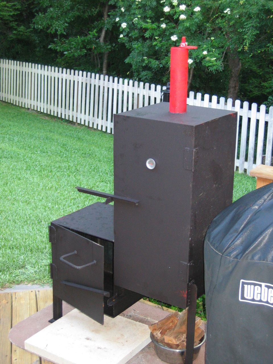 Black smoker with red chimney and open door, next to a Weber grill cover