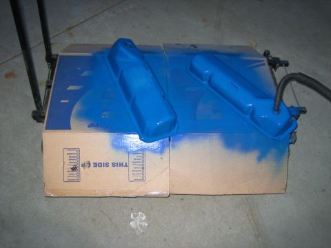 1979 Ford F-150 - Two blue painted valve covers are drying on top of cardboard boxes, with overspray visible on the boxes and the floor.