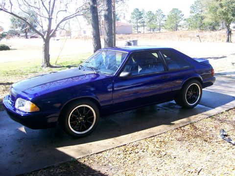 1991 Ford Mustang - A blue 1991 Ford Mustang coupe is parked on a concrete driveway with trees and a grassy area in the background.