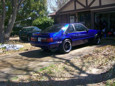 1991 Ford Mustang - A blue 1991 Ford Mustang is parked in front of a house, with another car visible in the background.