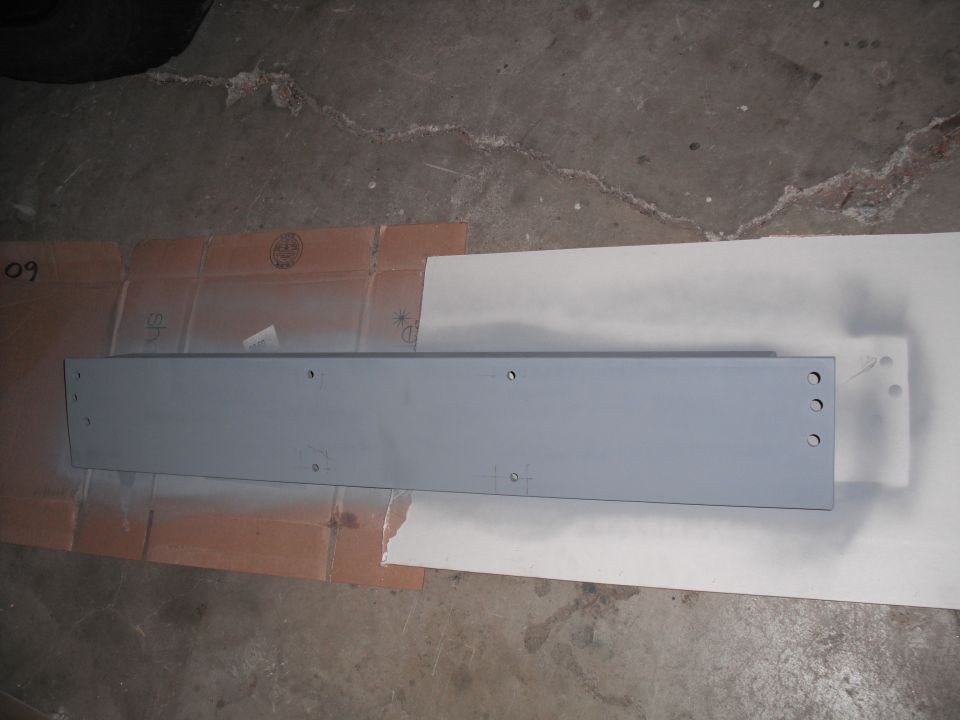 Gray painted metal piece with holes drilled in it, sitting on cardboard and paper