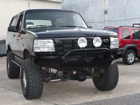 1995 Ford Bronco - This black 1995 Ford Bronco features an aftermarket bumper and lights, with a red SUV visible in the background.