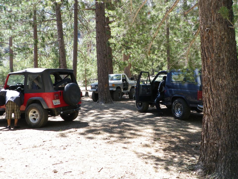 Several off-road vehicles parked in a forest clearing. A red Jeep Wrangler is in the foreground, with a white pickup truck and a dark SUV in the background