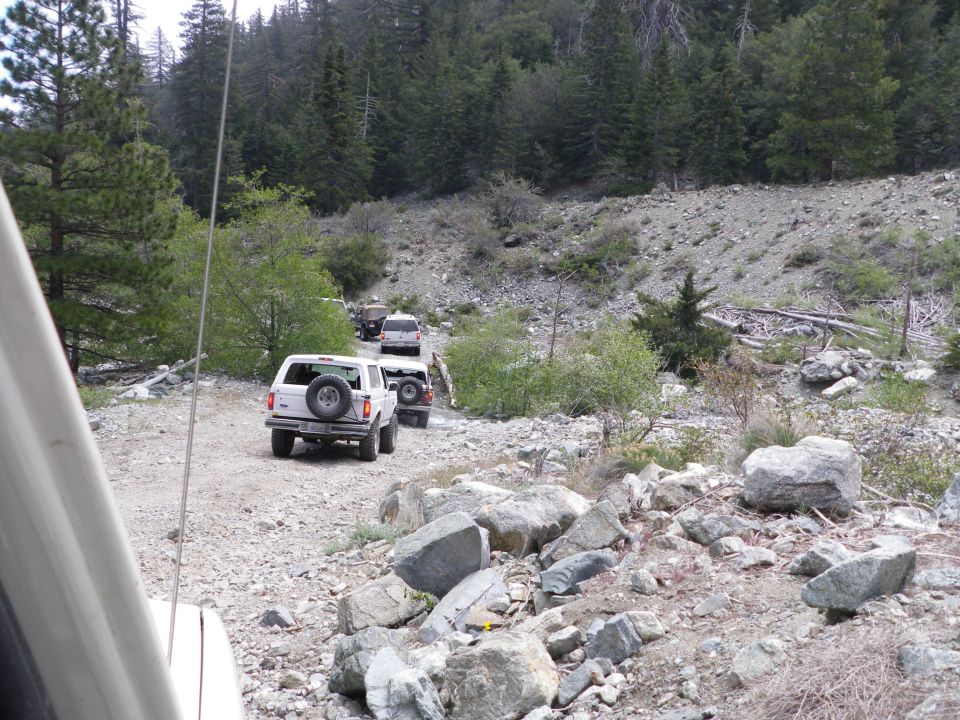 White 1991 Ford Bronco with spare tire on rear door, part of a convoy on a dirt road