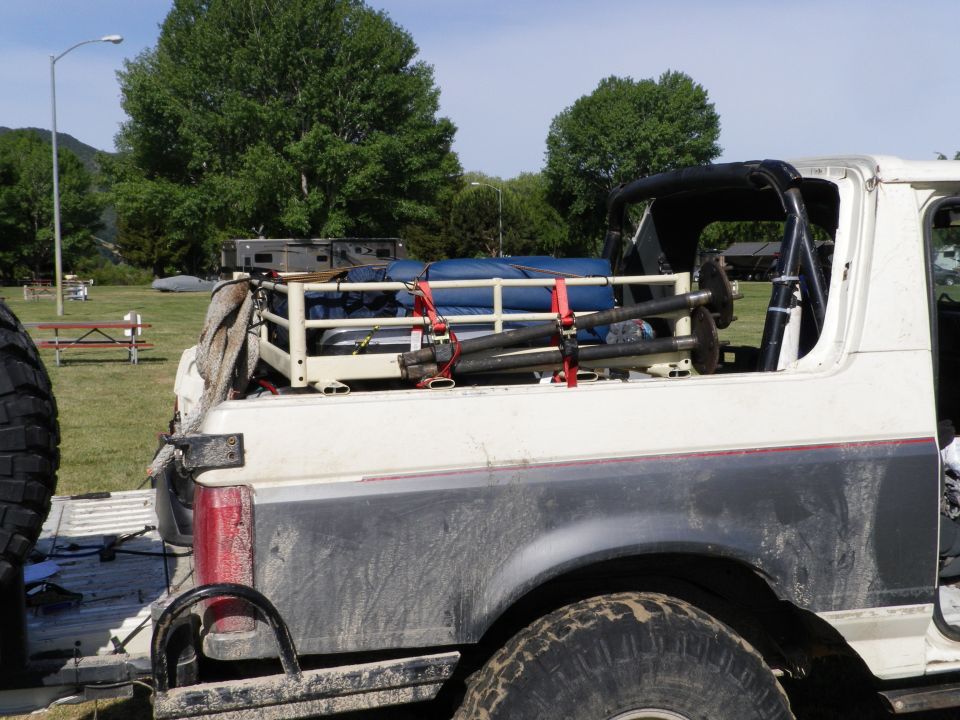 White Ford Bronco with a cargo rack loaded with blue tarps and poles, seen from the rear quarter