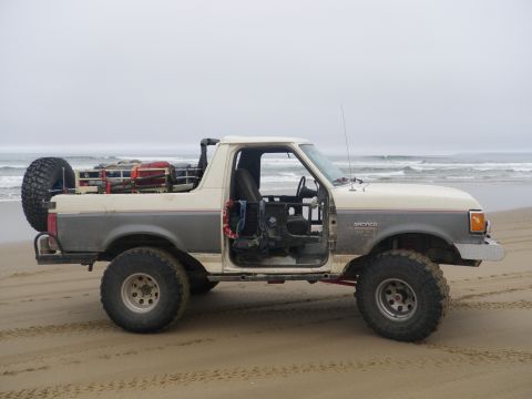 1991 Ford Bronco - Driver-side profile of a two-tone white-over-gray Ford Bronco on a foggy beach with surf breaking behind it, stripped to a doorless, toples…