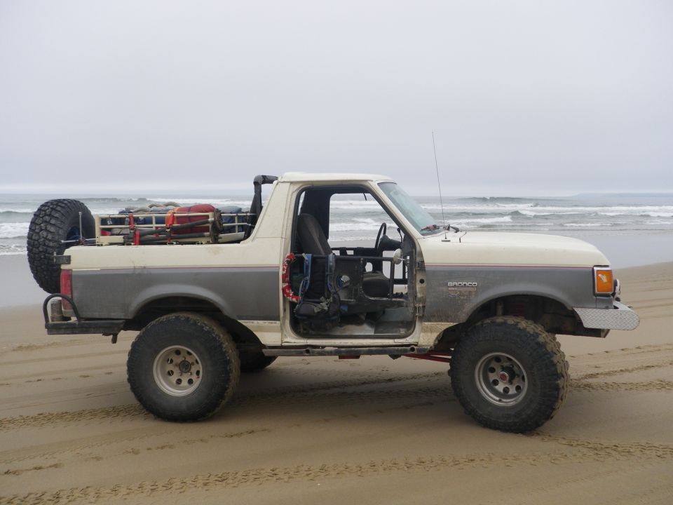 Doorless white and gray Ford Bronco with roll cage and spare tire parked on a foggy beach