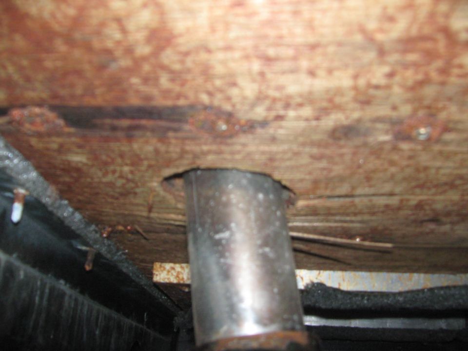 Metal pipe emerging from rusted wooden floor panel with visible screws and dark undercarriage