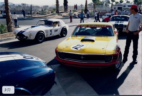 1973 Ford Mustang - A yellow 1970 Ford Mustang and a white Cobra are parked on a sunny day, with spectators in the background.