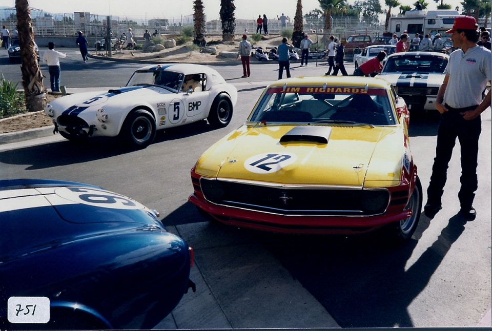 Yellow 1970 Ford Mustang with red trim and number 12 on hood, parked next to a white Cobra with number 5. Another blue Cobra is partially visible