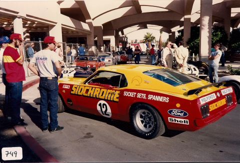 1973 Ford Mustang - A vintage racing car with prominent sponsor decals is parked outside a building, with several people around.