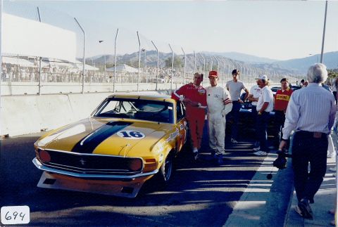 1973 Ford Mustang - A group of people stand around a vintage race car at a track, with spectators in the background.
