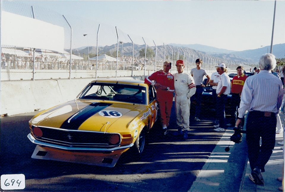 Yellow race car with black stripes and number 16 on hood, drivers in racing suits standing nearby