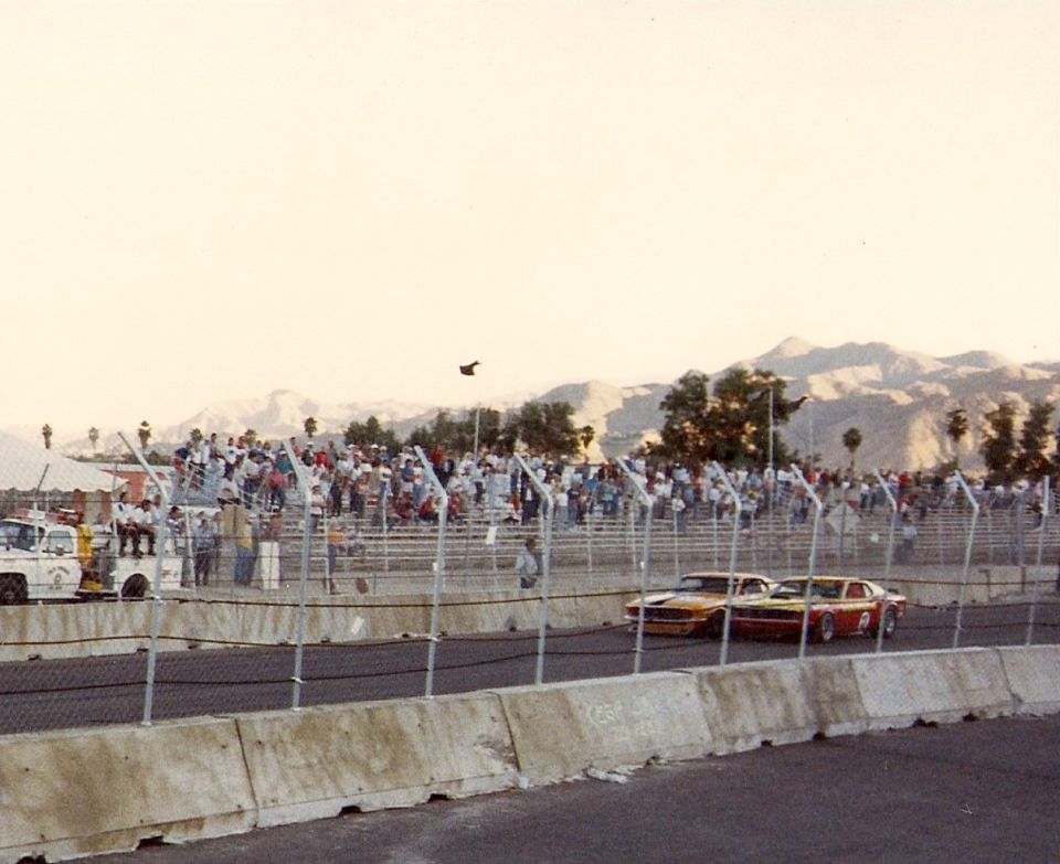 Two race cars, one orange and one red, on a track with spectators in the background