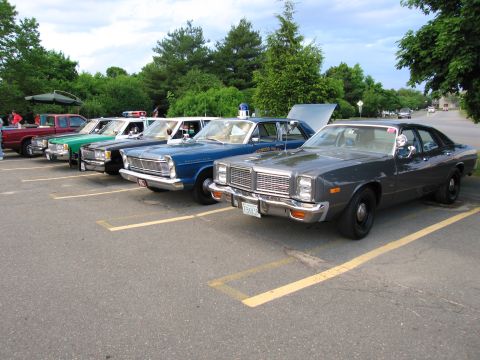 1991 Ford LTD Crown Victoria - row of vintage police sedans angled in a striped lot, led by a dark gray-blue Dodge cruiser with hood up.