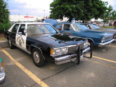 1991 Ford LTD Crown Victoria - black-and-white highway-patrol Ford sedan with star door emblem parked beside a blue vintage Westwood Police cruiser.